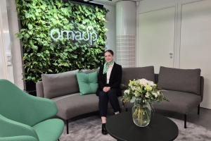 Kuvassa sohvalla Paula Hautamäki, taustalla viherseinä, jossa OmaSp:n logo valkoisella.