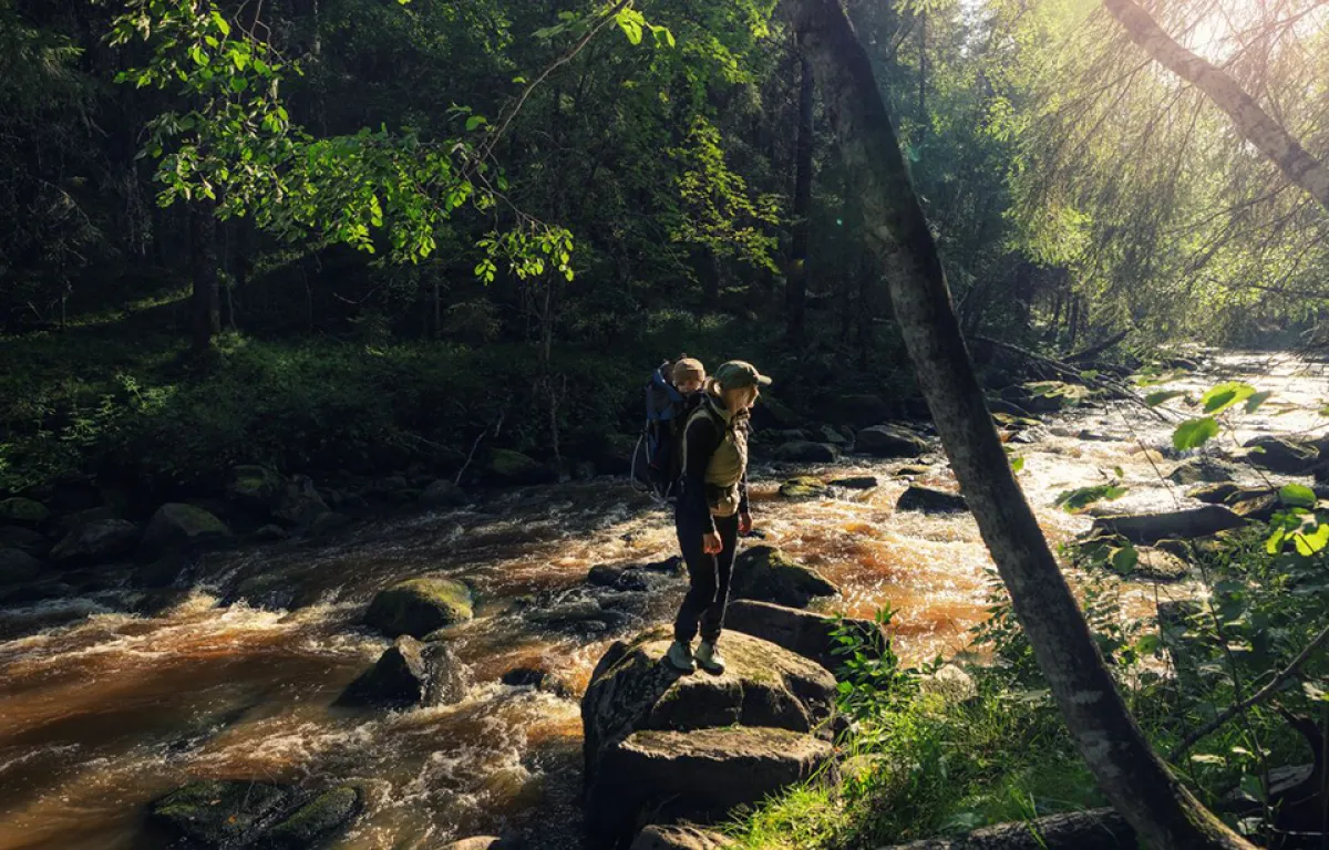 Nainen lapsi selässä patikoimassa aurinkoisella metsäpurolla.